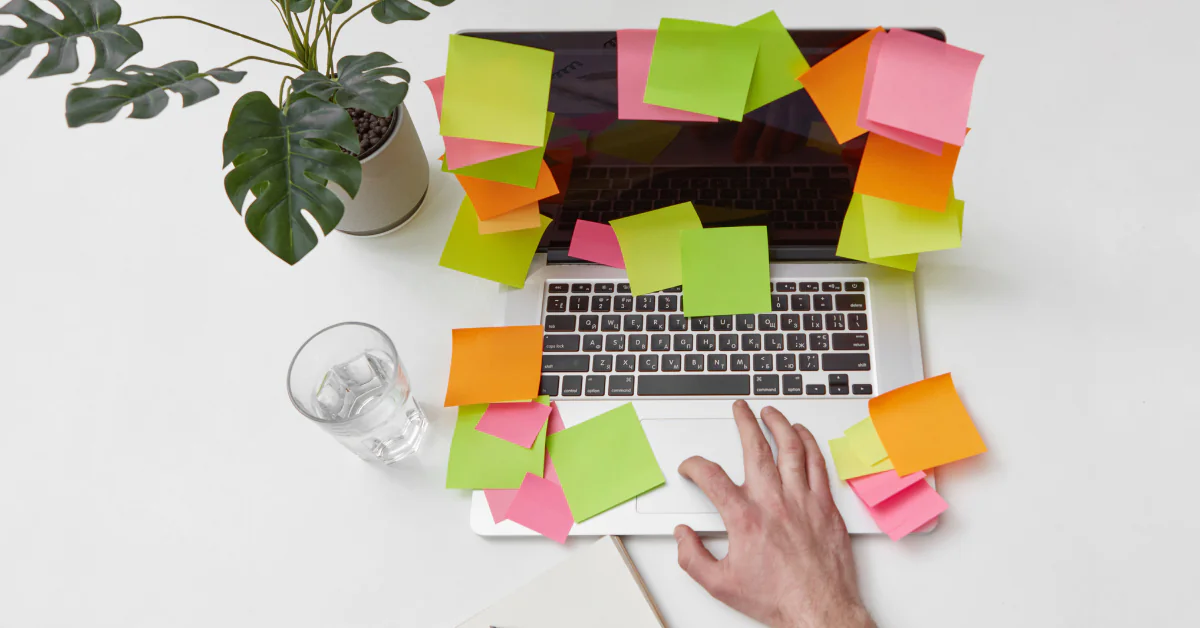 Computador com post-its coloridos ao redor do teclado, mesa organizada com planta e copo, representando estratégia prática para TDAH e procrastinação